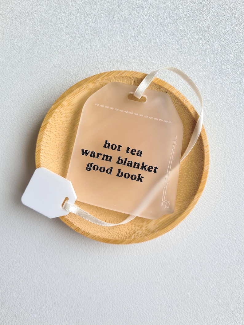 Puede incluir: Un posavasos de madera marr&oacute;n claro sostiene una etiqueta transl&uacute;cida en forma de bolsita de t&eacute; con las palabras "hot tea, warm blanket, good book" impresas en negro. Se adjuntan una cinta y una etiqueta blancas. El posavasos est&aacute; sobre una superficie blanca.