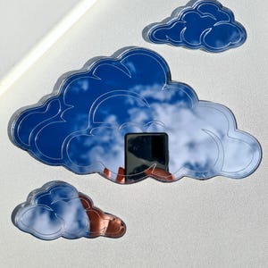 May include: Cloud-shaped wall decor in three sizes. The largest cloud reflects a blue sky with white clouds. The other two clouds are smaller, one blue and one with a copper-colored reflection. The decor is mounted on a white surface.