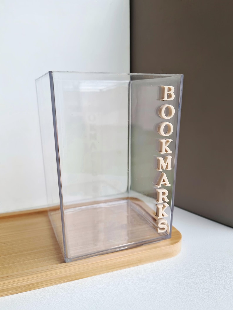 May include: A clear plastic container with the word "BOOKMARKS" spelled out in beige letters on the side. The container is sitting on a wooden surface.