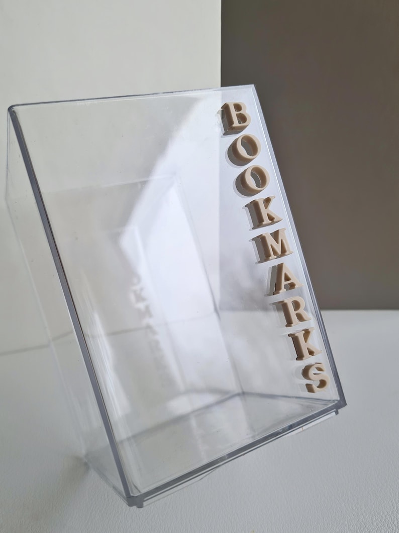 May include: A clear plastic container with the word "BOOKMARKS" spelled out in beige letters on the side.