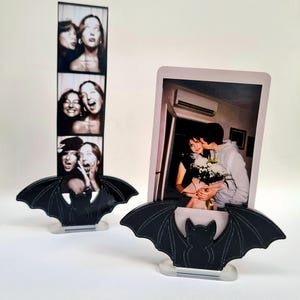 May include: Two photo frames with black bat-shaped bases. One frame holds a strip of black and white photos of women, the other holds a color photo of a couple. The frames are on a white surface.