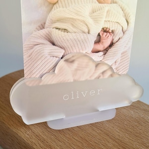 May include: A frosted acrylic cloud-shaped photo holder with the name "oliver" etched in white. The holder displays a photo of a baby wrapped in a cream-coloured blanket. The holder is on a wooden surface.