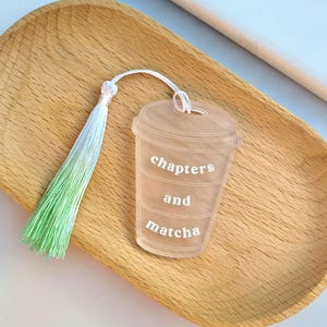 May include: A clear acrylic bookmark shaped like a coffee cup, with the words "chapters and matcha" in white. It has a white cord and a green and white tassel. The bookmark is on a wooden tray.