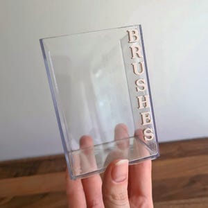 May include: A clear acrylic makeup brush holder with the word "BRUSHES" in light brown letters on the side. The holder is rectangular and held by a hand with a gold ring.