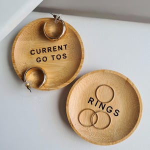 May include: Two wooden ring dishes. One dish is labelled "CURRENT GO TOS" and holds a pair of gold hoop earrings. The other dish is labelled "RINGS" and holds three gold rings.