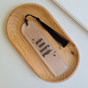 May include: A clear bookmark with a black tassel and the text "Just one more page..." printed on it. The bookmark is sitting on a wooden tray.