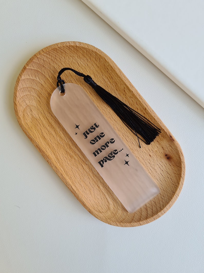 May include: A clear bookmark with a black tassel and the text "Just one more page..." printed on it. The bookmark is sitting on a wooden tray.