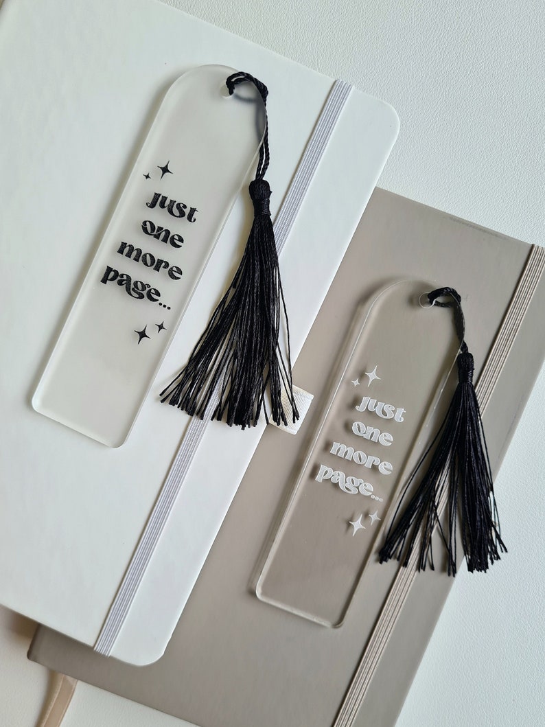 May include: Two clear acrylic bookmarks with black tassels. The bookmarks have the text "Just one more page..." in white lettering with small star accents.