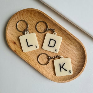 Może przedstawiać: Trzy białe plastikowe breloki w kształcie płytek do gry w Scrabble z czarnymi literami. Płytki tworzą słowo "LDK" i mają odpowiadające im wartości punktowe 1, 2 i 5.