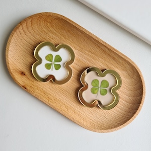 May include: Two gold-framed four-leaf clover charms, one with a white background and one with a light pink background. The charms are on a wooden tray.