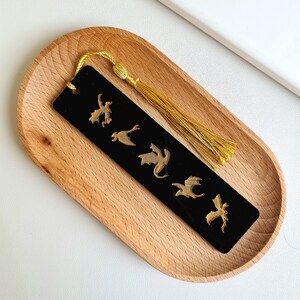 K&ouml;nnte beinhalten: Schwarzes Lesezeichen mit einem goldenen Quasten und einem Design aus f&uuml;nf goldenen Drachen. Das Lesezeichen befindet sich auf einem Holztablett.