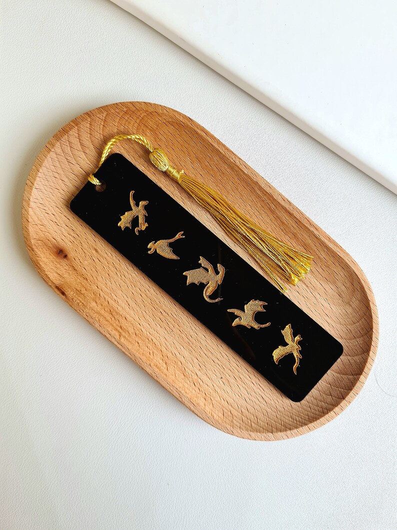 K&ouml;nnte beinhalten: Schwarzes Lesezeichen mit einem goldenen Quasten und einem Design aus f&uuml;nf goldenen Drachen. Das Lesezeichen befindet sich auf einem Holztablett.