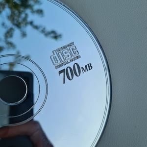 May include: A silver compact disc with the text "COMPACT disc DIGITAL AUDIO" and "700MB" printed on the surface. The disc reflects the surrounding environment, including trees and a hand holding a phone.