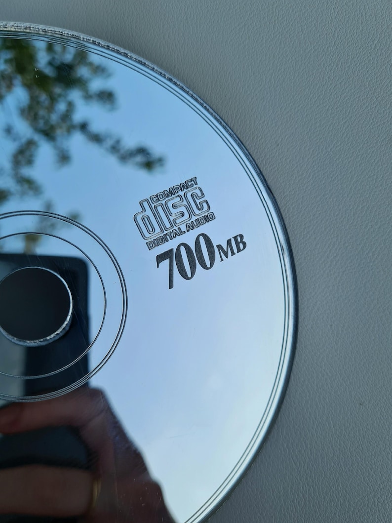 May include: A silver compact disc with the text "COMPACT disc DIGITAL AUDIO" and "700MB" printed on the surface. The disc reflects the surrounding environment, including trees and a hand holding a phone.