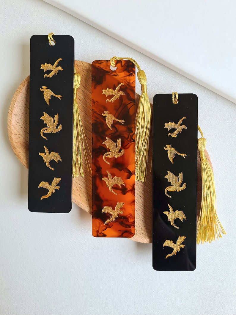 K&ouml;nnte beinhalten: Drei Lesezeichen mit goldenen Drachen-Silhouetten auf schwarzem und braunem Hintergrund. Jedes Lesezeichen hat einen goldenen Quasten.