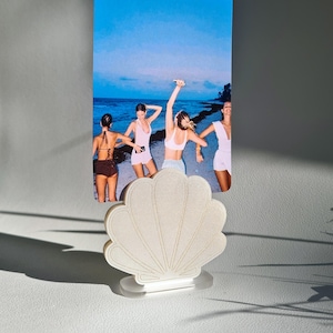 Peut inclure: Un porte-photo en forme de coquillage, avec une base transparente, tenant une photo rectangulaire de quatre personnes sur une plage. Le porte-photo est blanc cassé avec des lignes dorées. La photo montre des personnes en maillots de bain et en tenues décontractées.