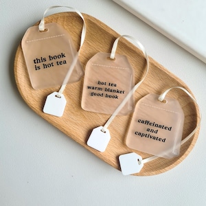Puede incluir: Tres marcap&aacute;ginas con forma de bolsita de t&eacute; con etiquetas y cintas blancas sobre una bandeja de madera. Cada marcap&aacute;ginas tiene una frase diferente: "this book is hot tea", "hot tea warm blanket good book", y "caffeinated and captivated".