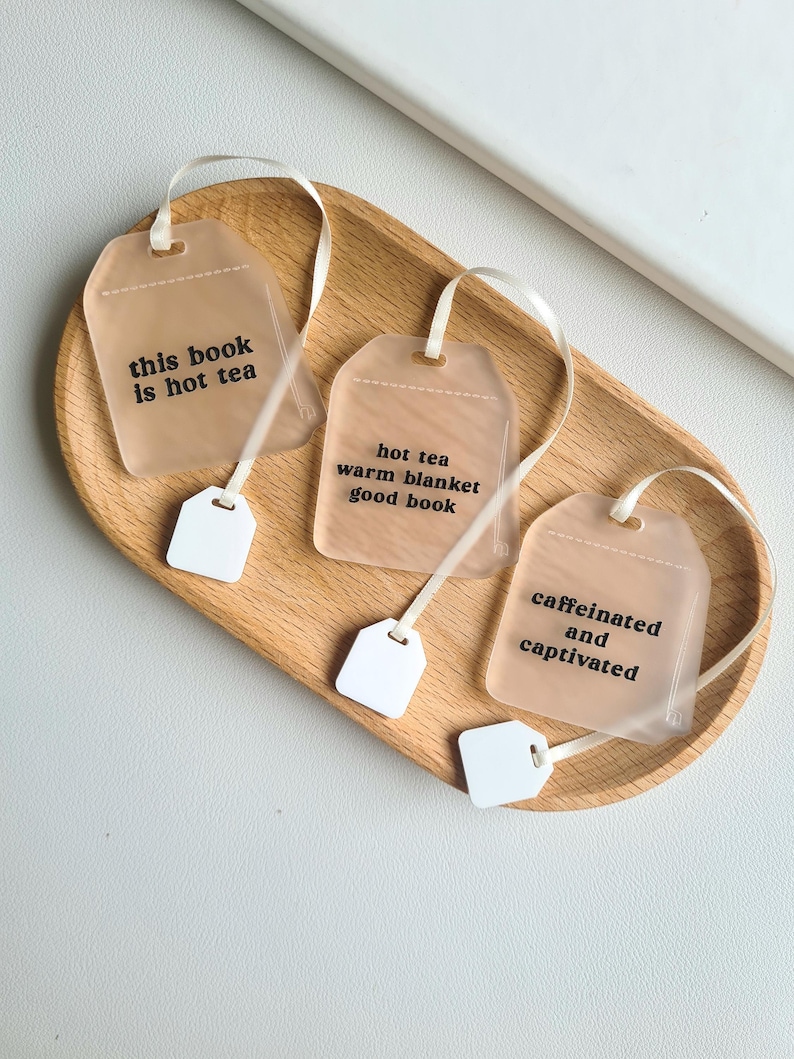 Puede incluir: Tres marcap&aacute;ginas con forma de bolsita de t&eacute; con etiquetas y cintas blancas sobre una bandeja de madera. Cada marcap&aacute;ginas tiene una frase diferente: "this book is hot tea", "hot tea warm blanket good book", y "caffeinated and captivated".