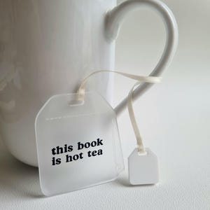 Puede incluir: Una taza de cer&aacute;mica blanca con un asa curvada. Un objeto transparente en forma de etiqueta con el texto "this book is hot tea" est&aacute; sujeto a la taza con una cinta color crema. Tambi&eacute;n hay una peque&ntilde;a etiqueta blanca sujeta a la cinta.