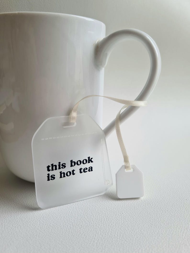 Puede incluir: Una taza de cer&aacute;mica blanca con un asa curvada. Un objeto transparente en forma de etiqueta con el texto "this book is hot tea" est&aacute; sujeto a la taza con una cinta color crema. Tambi&eacute;n hay una peque&ntilde;a etiqueta blanca sujeta a la cinta.