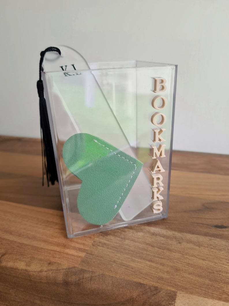 May include: A clear acrylic container with the word "BOOKMARKS" spelled out in wooden letters. Inside the container are two bookmarks, one is a plain white bookmark with a black tassel and the other is a green heart-shaped bookmark.