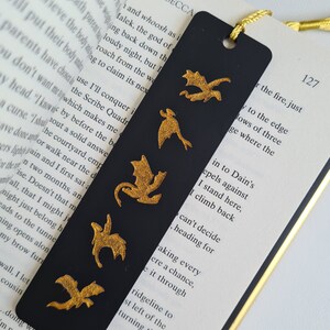 K&ouml;nnte beinhalten: Schwarzes Lesezeichen mit f&uuml;nf goldenen Drachen-Silhouetten. Das Lesezeichen liegt auf einer Buchseite mit dem sichtbaren Text "REBECCA" und "127".