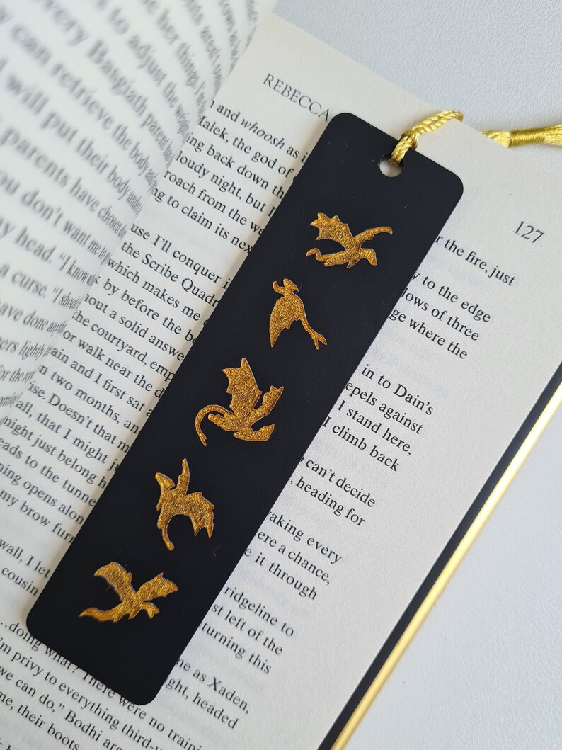 K&ouml;nnte beinhalten: Schwarzes Lesezeichen mit f&uuml;nf goldenen Drachen-Silhouetten. Das Lesezeichen liegt auf einer Buchseite mit dem sichtbaren Text "REBECCA" und "127".