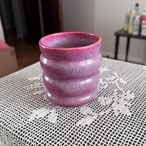 May include: A cylindrical ceramic cup featuring a ribbed, three-tiered design. The exterior is finished with a mottled glaze that transitions from a deep magenta at the rim to a lighter, soft lavender-purple hue across the body of the vessel.