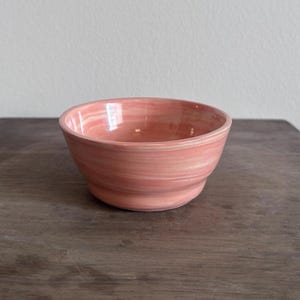 May include: A small, round ceramic bowl featuring a marbled glaze with swirling shades of light and dark pink. The bowl has a smooth, glossy finish and a slightly flared rim, with a subtle horizontal ridge encircling the lower portion of the body.