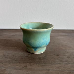 May include: A small, flared ceramic cup featuring a smooth, glossy glaze in shades of seafoam green and turquoise. The vessel has a rounded body that tapers toward a narrow base, with subtle variations in color intensity across the surface.