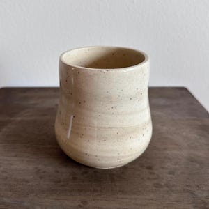 May include: A cream-colored ceramic mug with a speckled finish and a rounded, slightly tapered body. The surface features subtle horizontal ridges along the sides, creating a textured appearance. The interior is smooth and matches the exterior color.