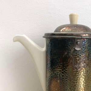 Könnte beinhalten: Eine silberne und weiße Teekanne mit gehämmerter Metalloberfläche und einem weißen Porzellan-Ausguss und Griff.