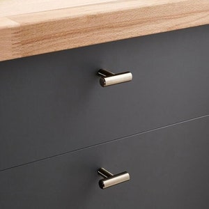 May include: A close-up shot of a dark grey cabinet with two drawers. Each drawer features a gold-coloured, cylindrical handle. The cabinet top is made of light-coloured wood.
