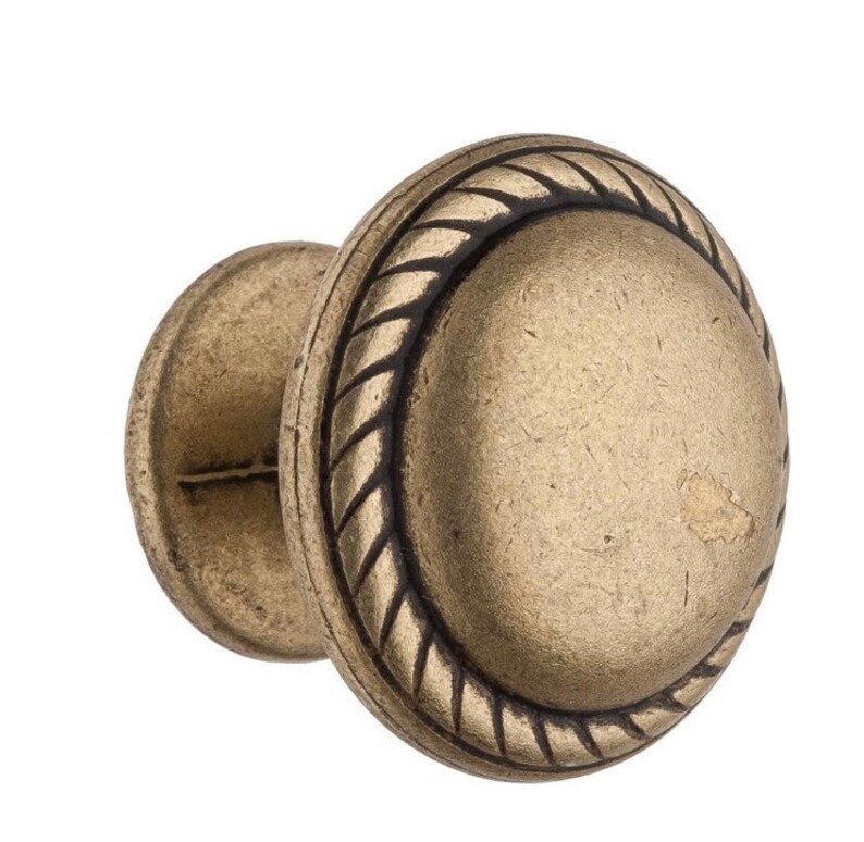 Może przedstawiać: Złoty gałka do szafki z wzorem sznurka wok&oacute;ł krawędzi. Gałka ma okrągły kształt i lekko wypukły środek.