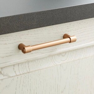 May include: A bronze-coloured cabinet pull with a cylindrical bar and rounded ends. The hardware is mounted on a white wooden cabinet with visible wood grain. The pull is approximately 15 cm long.