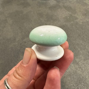 May include: A ceramic mushroom-shaped cabinet knob. The base is white, and the top is a gradient of green and white. The knob has a glossy finish. The knob is held in a hand.