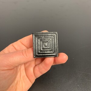 May include: A black ceramic tile with a concentric square design.