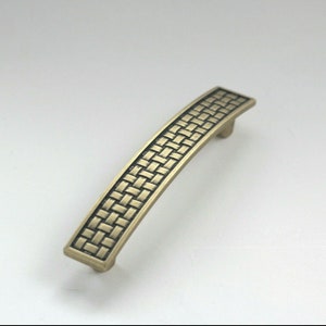 May include: A bronze-finished cabinet pull with a woven pattern. The curved handle has a rectangular shape and a textured surface. This furniture hardware is designed for cabinets and drawers, with a length of approximately 15 cm.
