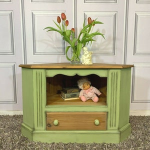 May include: A green corner TV stand with a shelf and a drawer. The stand is painted in a distressed style and has a wooden top. A small doll is sitting on the shelf.