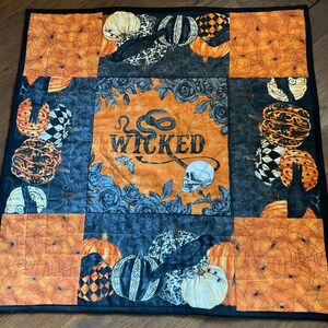 May include: A Halloween-themed quilted table runner featuring pumpkins, crows, and spiderwebs in orange, black, and white. The center panel has the word "WICKED" with a skull and snake design. The runner is square with a black border.