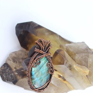 May include: A blue labradorite gemstone pendant wrapped in copper wire. The pendant is displayed on a bed of brown and clear quartz crystals.
