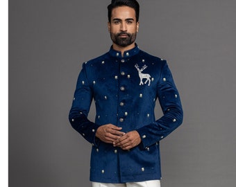 Könnte beinhalten: Marineblaue Samtjacke mit goldenen Knöpfen und einem Hirsch-Emblem, kombiniert mit einer weißen Hose und blauen Schuhen. Die Jacke hat kleine goldene Verzierungen. Das Outfit ist formell und elegant.