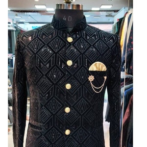 May include: A black velvet blazer with a geometric pattern and gold buttons. The blazer has a mandarin collar and a pocket on the left side with a gold chain and a decorative pin.