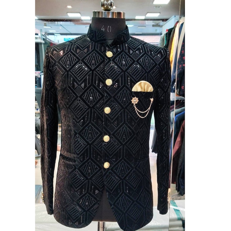 May include: A black velvet blazer with a geometric pattern and gold buttons. The blazer has a mandarin collar and a pocket on the left side with a gold chain and a decorative pin.