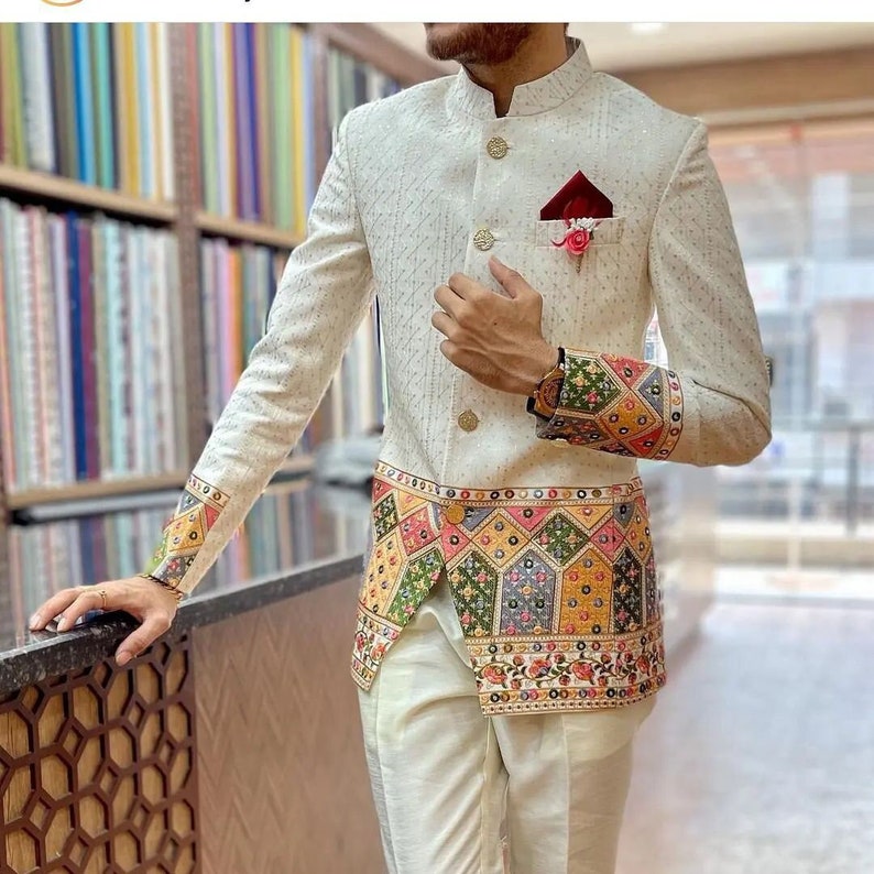 May include: A white blazer with a mandarin collar and gold buttons. The blazer has a colorful patterned trim around the bottom and cuffs. The blazer is worn over a pair of white pants.