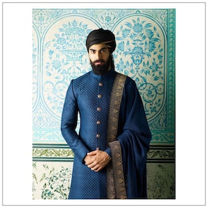 May include: A man wearing a blue quilted sherwani with a gold and black embroidered shawl. He is wearing a black turban and has a beard.