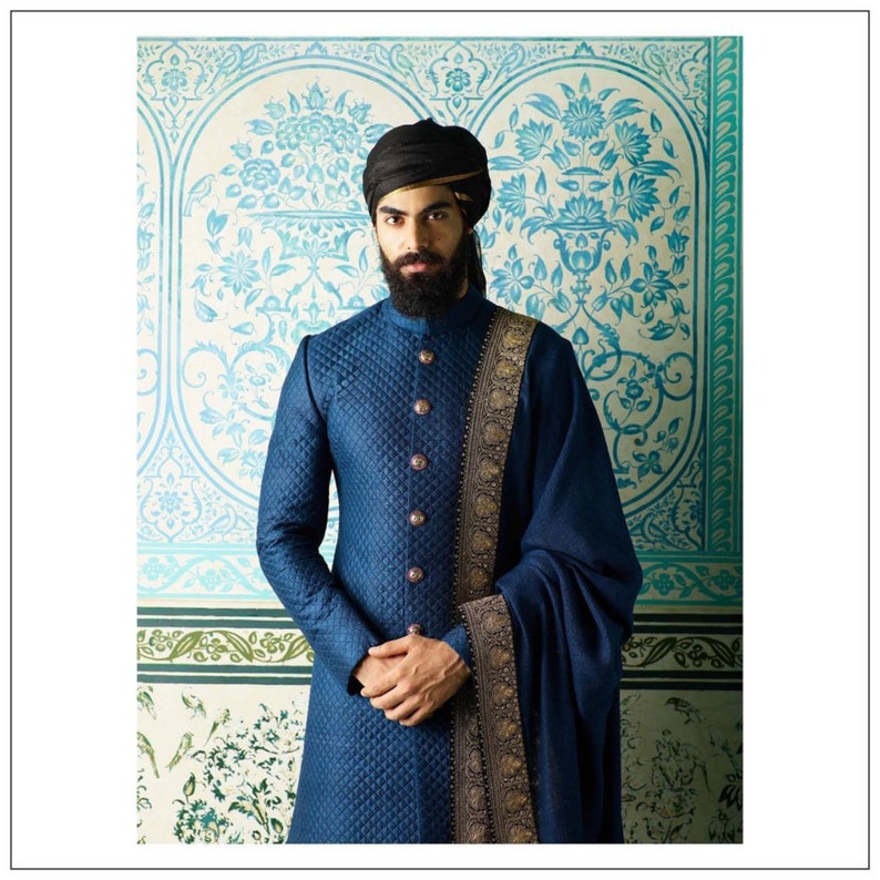 May include: A man wearing a blue quilted sherwani with a gold and black embroidered shawl. He is wearing a black turban and has a beard.