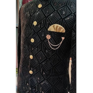 May include: A black velvet jacket with a geometric pattern and gold buttons. The jacket has a pocket on the left side with a gold chain and a decorative pin.