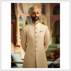 May include: A man wearing a white quilted long-sleeved jacket with a mandarin collar and gold buttons. He is wearing a patterned turban and has a beard.