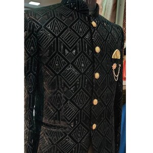 May include: A black velvet blazer with a geometric pattern and gold buttons. The blazer has a mandarin collar and a pocket on the left side. A gold chain with a decorative pin is attached to the pocket.